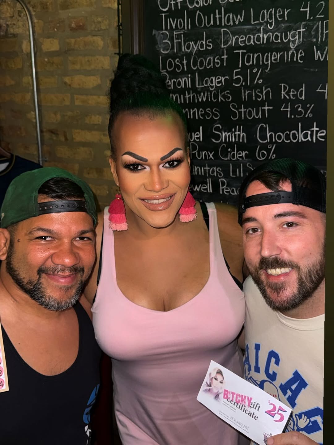 Bitchy Bingo host Batty Davis with two smiling winners holding a $25 gift certificate in front of the craft beer chalkboard